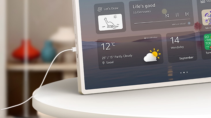 LG StanbyME 2 Max LX6 Beweglicher Bildschirm mit USB-C-Kabel wird auf einer Tabelle mit einem Kabel am Anschluss angezeigt, das eine Heim-Widget-Schnittstelle zeigt, die gleichzeitiges Laden und die Gerätekonnektivität hervorhebt.